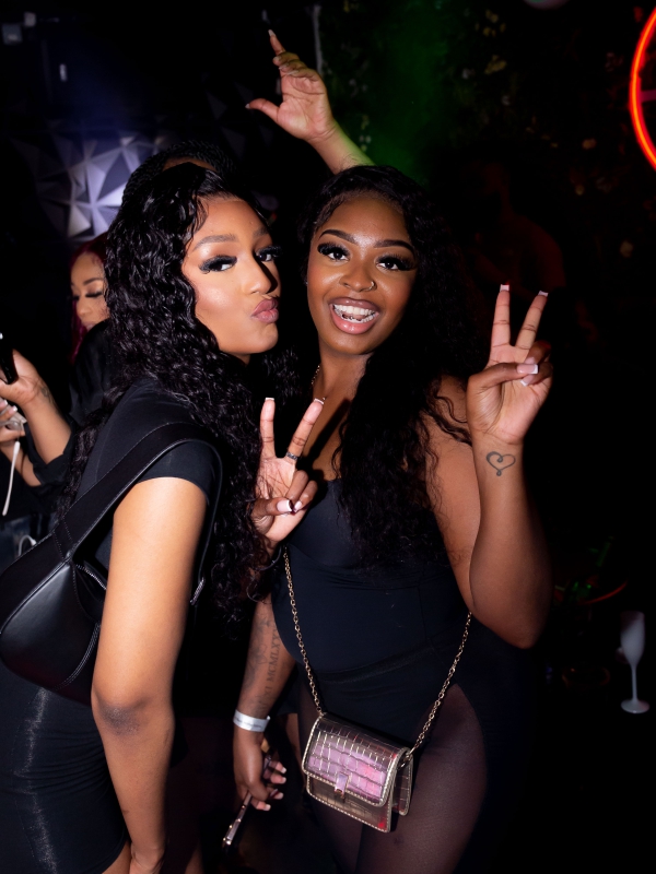 Two Women Posing Together And Making Peace Signs At A Dimly Lit Evening Party.